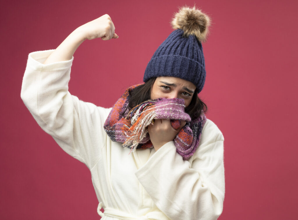weak young ill woman wearing robe winter hat scarf covering mouth with scarf looking front doing strong gesture isolated pink wall